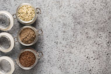 Kenevir taneleri, kahverengi keten tohumu ve gri taş masadaki cam kavanozda keten tohumu. Üst Manzara. Metin için boşluk.