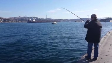 Video taken on 8th March 2020:Back of man fishing alone at a beautiful spot on a bright sunny day and People walking along promenade of Istanbul resort city