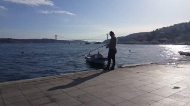 Video taken on 8th March 2020:Back of man fishing alone at a beautiful spot on a bright sunny day and People walking along promenade of Istanbul resort city
