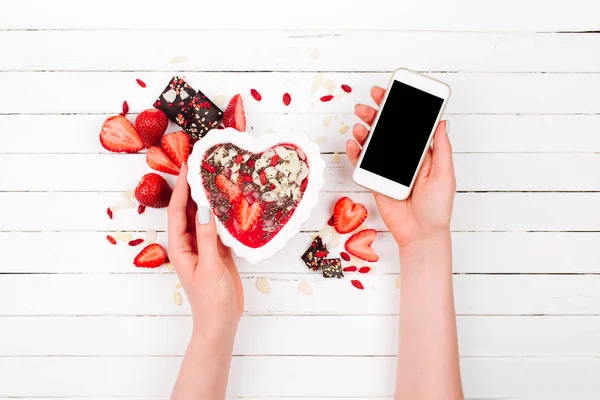 Tangan wanita memegang smoothie stroberi dalam mangkuk dalam bentuk hati dan smartphone. Hari Valentine. Sarapan hari libur — Stok Foto