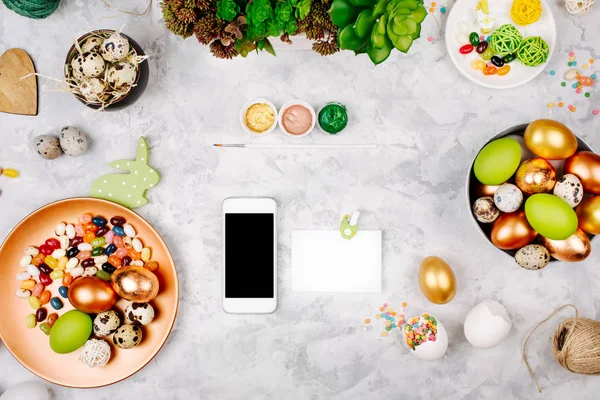 Smartphone dan kartu kosong dengan latar belakang dekorasi easter. Dicat telur di nampan, permen, bunga dan cat — Stok Foto