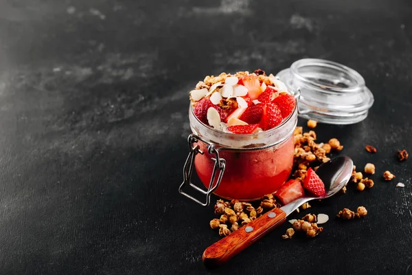 Café da manhã saudável, smoothie de baga em um frasco de vidro coberto com morango, flocos de amêndoa e granola no fundo preto — Fotografia de Stock