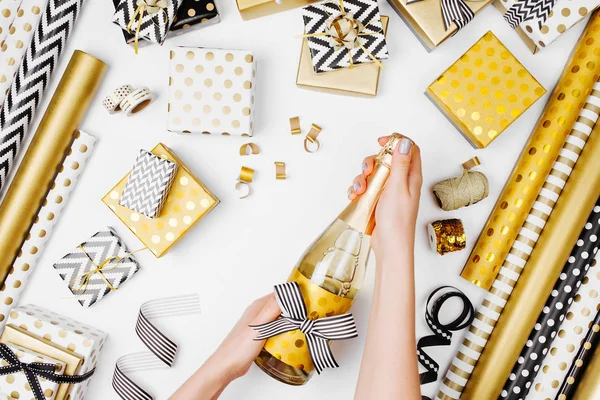 female hands opening champagne bottle on the table with flat lay of golden presents and wrapping paper rolls
