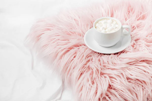 Cup of cacao with marshmellow on pink furry pillow on bed