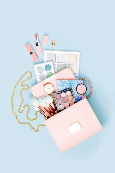 Cosmetic products flowing from makeup bag on pastel blue background.