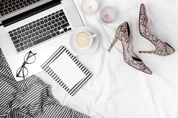 Fashion blogger workspace with laptop, snake shoes, glasses, coffee, candles and notebook in bed.