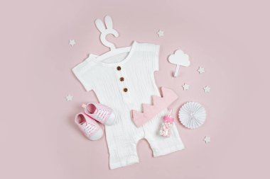 Set of baby clothes and accessories on pink background. White bodysuit on cute hanger with bunny ears and baby shoes, cotton crown and toys. Fashion newborn clothes. Flat lay, top view