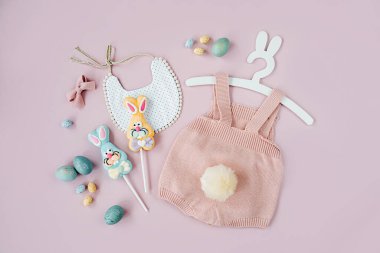 Cute baby costume Easter bunny. Knitted bodysuit with fluffy tail bunny, Easter eggs and candy on pink background. Holiday concept. Flat lay, top view