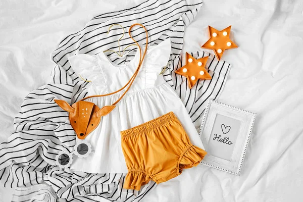 stock image White dress, orange shorts with kids handbag and sunglasses. Set of  baby clothes and accessories for summer vacation on  bed. Fashion childs outfit. Flat lay, top view