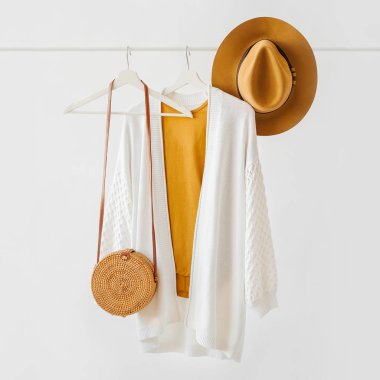 White knitted jumper on hanger with brown hat and bamboo bag on white background. Elegant  fashion outfit. Spring wardrobe. Minimal concept.