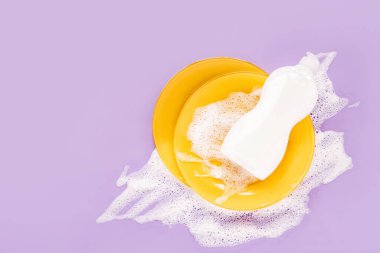 Yellow plates and bottle with washing agent on soapy foam background. Washing dishes concept. Flat lay, Top view.