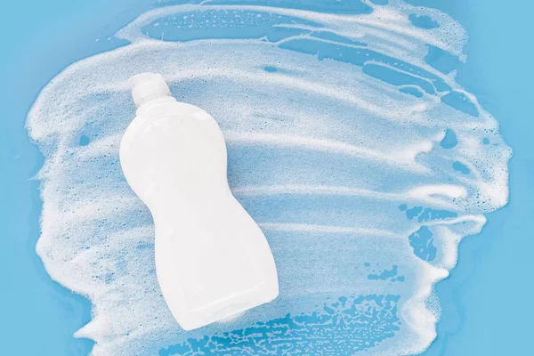 Bottle with washing agent with white soapy foam on a blue background. Cleaning/Washing concept. Flat lay, Top view.