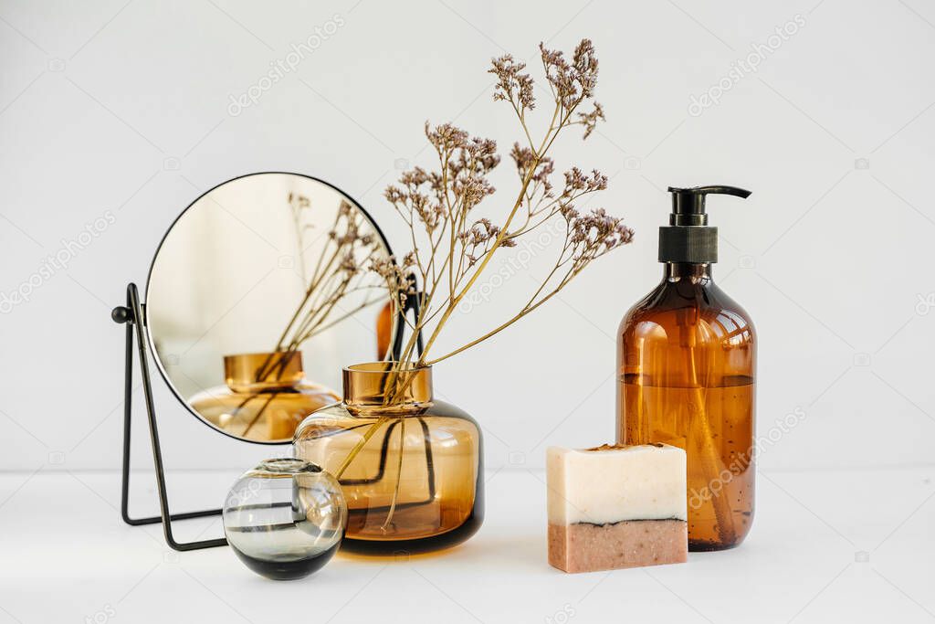 Branch flowers in a glass vase, mirror,  shampoo bottles and soap on white table. Decor for interior. Stylish decoration for home.