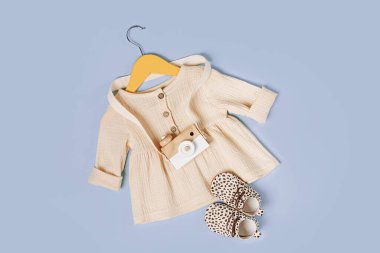 White dress on hanger with toy camera and shoes. Set of baby clothes and accessories for spring, autumn or summer on light blue background. Fashion kids outfit. Flat lay, top view