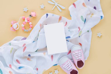 Boş kartlı muslin kundak battaniyesi ve bebek terlikleri, sarı arka planda oyuncaklar. Mesajı hazırla. Yeni doğmuş aksesuarlar. Düz yatış, üst görünüm 
