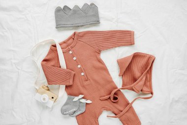 Beyaz yatakta bebek tulumu, şapka, çorap ve oyuncak seti. Cinsiyet ayrımı olmayan bebek kıyafetleri ve aksesuarlar. Düz yatış, üst görünüm