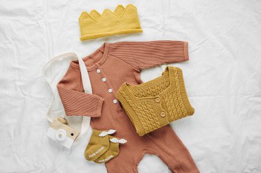 Beyaz yatakta bebek tulumu, şapka, çorap ve oyuncak seti. Cinsiyet ayrımı olmayan bebek kıyafetleri ve aksesuarlar. Düz yatış, üst görünüm