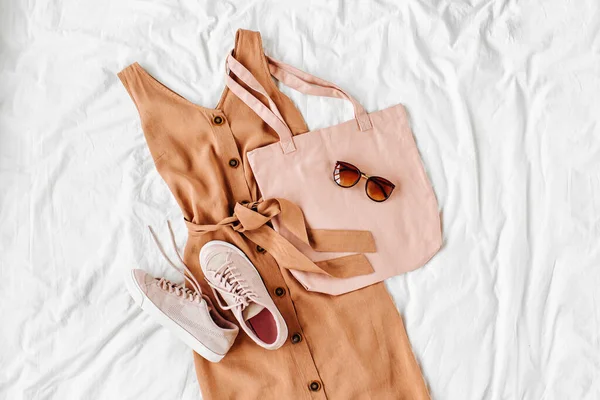 Beige dress with eco bag, sneakers and accessories on white bed. Women\'s stylish autumn or summer outfit. Trendy clothes. Flat lay, top view.