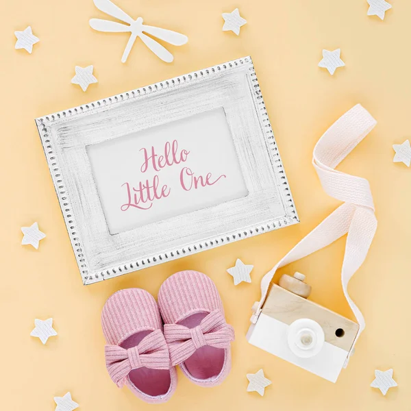 Photo frame with card Hello little one and baby slippers, toys on yellow background.  Set of  newborn accessories for girl. Flat lay, top view