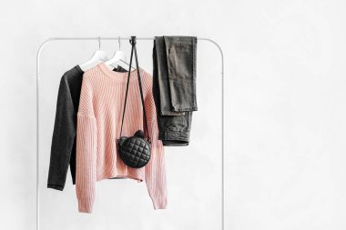 Female clothes in pastel pink and gray color on hanger on white background.  Jumper, shirt, jeans and bag. Spring/autumn outfit. Minimal concept.