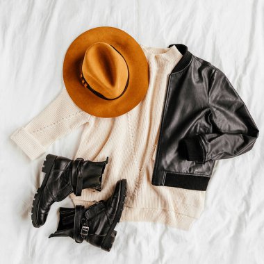 Deri ceket ve beyaz yatağın üzerinde kahverengi şapkalı ve çizmeli sıcak kazak. Kadınların şık sonbahar ya da kış kıyafetleri. Moda kıyafet kolajı. Düz yatış, üst görünüm. 