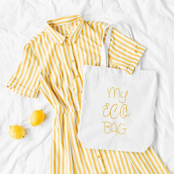 Yellow dress with stripes with eco bag  on white bed. Women\'s stylish  summer outfit. Trendy clothes with white eco bag mockup. Flat lay, top view.