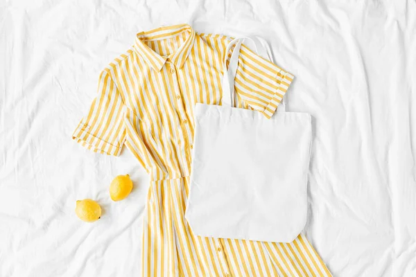 Yellow dress with stripes with eco bag  on white bed. Women\'s stylish  summer outfit. Trendy clothes with white eco bag mockup. Flat lay, top view.