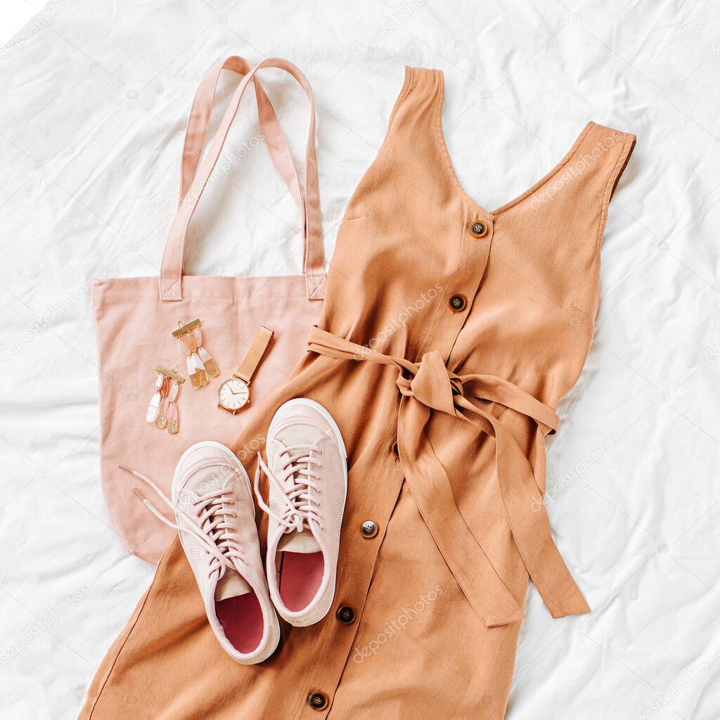 Beige dress with eco bag, sneakers and accessories on white bed. Women's stylish autumn or summer outfit. Trendy clothes. Flat lay, top view.