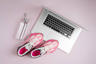 Laptop, spor ayakkabılar ve pembe arka planda su şişesi. Çevrimiçi Fitness programı. Ev antrenmanı. Üst manzara, düz uzanma