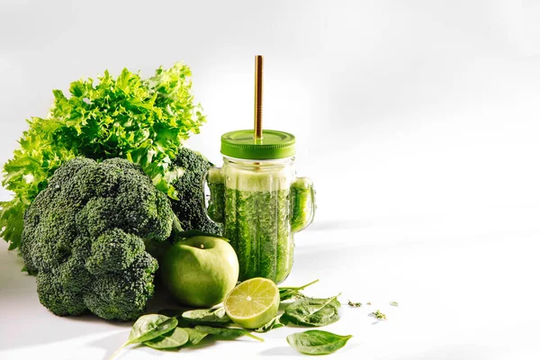 Green smoothie in cute glass jar shape of cactus with spinach and green fruits and vegetables on white table. Vegetarian food. Detox and diet concept.