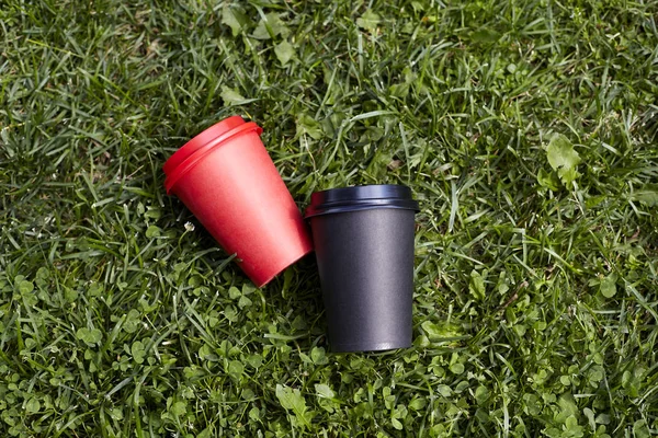 Two paper cups with coffee to take away, red and  black cup on green grass outdoor breakfast