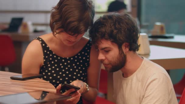 若いカップルは スマートフォンを見てコーヒーショップでいくつかのアプリについて話します — ストック動画