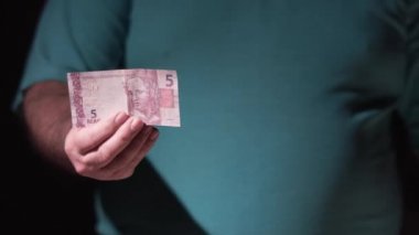 Şişman yaşlı bir adam, Brezilyalı kamera için 5 reais gösteriyor.