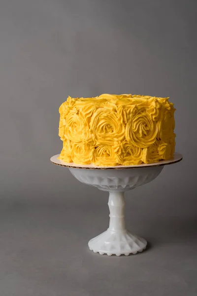 Yellow frosted cake with a rose design sitting on a white cake stand with gray background