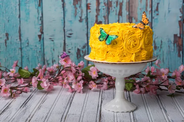 Yellow cake sitting on a cake stand.  Turquoise and white peeled paint background and table pink flowers and butterflies