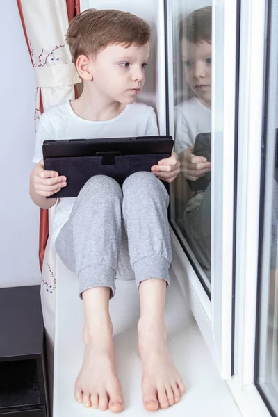 Kişisel izolasyon modunda ve karantina konseptinde uzaktan öğrenme. Yedi yaşında bir çocuk pencerenin yanında oturuyor ve elinde bir tablet tutuyor. Çevrimiçi okul, video konferans dersleri