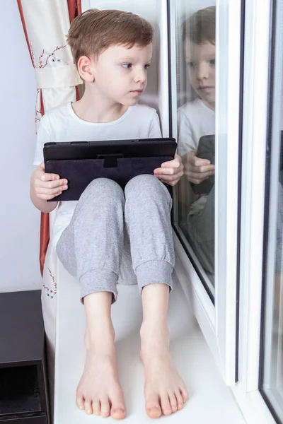 Afstandsonderwijs in zelfisolatiemodus en quarantaineconcept. Een zevenjarige jongen zit bij het raam en houdt een tablet in zijn handen. Online school, videovergaderlessen Rechtenvrije Stockafbeeldingen