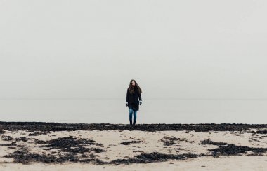 Genç kız siyah uzun ceket ve kot siyah deniz yosunu ile plaj boyunca denizden gider. Jurmala Letonya