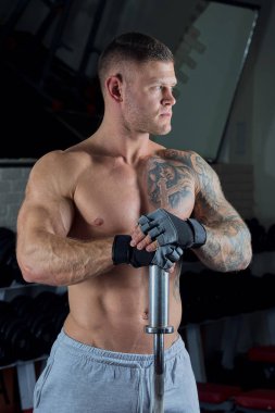   Kas gömleksiz adam mavi gözleri ve dövme halter eldiven ve bir spor salonu gri pantolon ile poz veriyor 