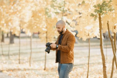 Deri ceketli, mavi gömlekli ve kot pantolonlu kel, vahşi bir fotoğrafçı öğleden sonra parktaki kameradan fotoğraf seçiyor.