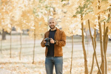 Deri ceketli, mavi gömlekli ve kot pantolonlu kel, vahşi fotoğrafçı kamerayı tutuyor ve öğleden sonra parkta gülümsüyor.