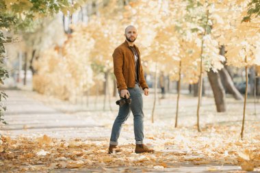 Deri ceketli, mavi gömlekli, kot pantolonlu ve Chelsea botlu kel bir fotoğrafçı kamerayı tutuyor ve öğleden sonra parkta bir mankeni bekliyor.