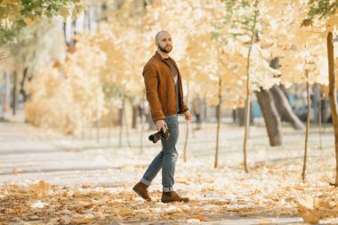 Süet deri ceketli, mavi gömlekli, kot pantolonlu ve Chelsea botlu kel bir fotoğrafçı kamerayı tutuyor ve öğleden sonra parkta poz veriyor.