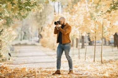 Süet deri ceketli, mavi gömlekli, kot pantolonlu ve Chelsea botlu kel bir fotoğrafçı güneşli bir öğleden sonra parktaki kamerada fotoğraf çekiyor.
