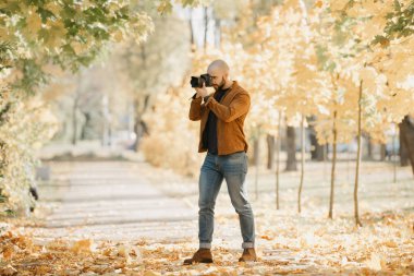 Bald stylish photographer with a beard in a suede leather jacket, blue shirt, jeans, and Chelsea boots takes photos on the camera in the park in the sunny afternoon clipart