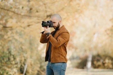 Süet deri ceketli, mavi gömlekli, kotlu, dijital kol saatli kel bir fotoğrafçı güneşli bir öğleden sonra ormanda fotoğraf çeker.