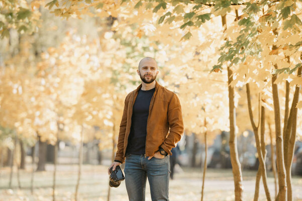 Bald brutal photographer with a beard in a suede leather jacket, blue shirt and jeans holds the camera and waits for a model in the park in the afternoon