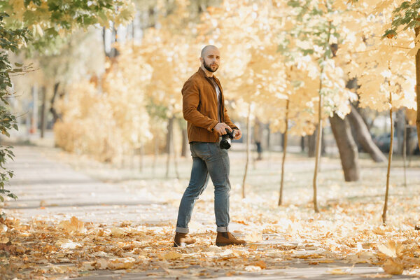 Лысый безжалостный фотограф с бородой в замшевой кожаной куртке, голубой рубашке, джинсах и ботинках Челси держит камеру и ждет модель в парке во второй половине дня
