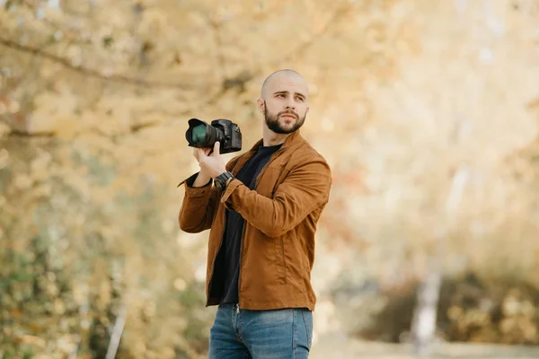 While bald stylish photographer with a beard in a suede leather jacket, blue shirt and jeans with digital wristwatch looks through photos in his camera, he drew attention to something in the forest in the sunny afternoon
