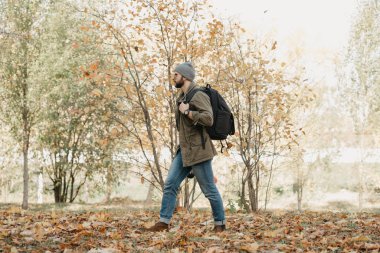 Aynalı gözlüklü, havacı güneş gözlüklü, zeytin askeri ceketli, kot pantolonlu, sırt çantalı ve kol saatli vahşi bir gezgin öğle vakti ormandan geçer. Bir foto muhabiri.